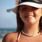 Purple, yellow, & black Bermuda Triangle heishi necklace on smiling model wearing hat on beach.